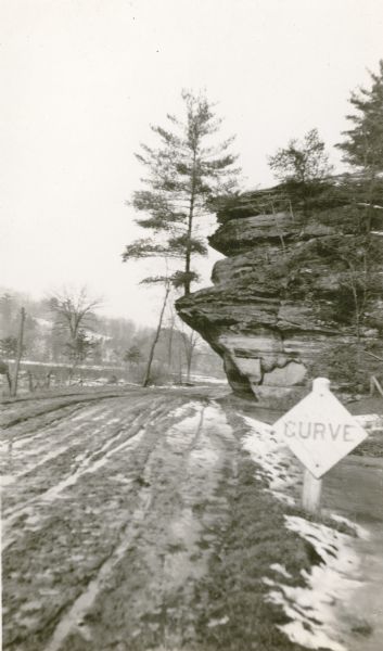 A single pine tree grows on the ledge of a bluff along Highway 80 near Hub City. A road sign warns of a curve. The road is rutted and there is patchy snow on the ground.