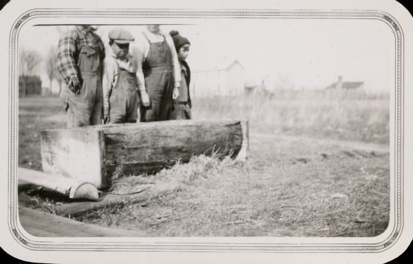 Three boys and a girl stand behind a wood trough, hewn from a log. The boys wear bib overalls. There is a two-story house and an outbuilding in the background. One of the boys is African American.