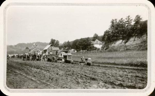 Men using horse-drawn and gasoline powered road equipment work on Highway 80 near Rockbridge. A wooded bluff rises steeply behind a farmhouse. Barns, a silo, and outbuildings stand closer to the road.