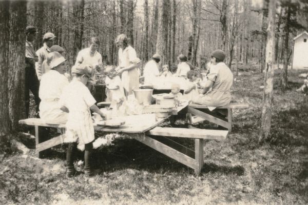 Children and adults enjoy a picnic in the wooded schoolyard at the Cloverland School, District No. 2. Pans of food have been placed on boards supported by benches. There is an outhouse in the background.