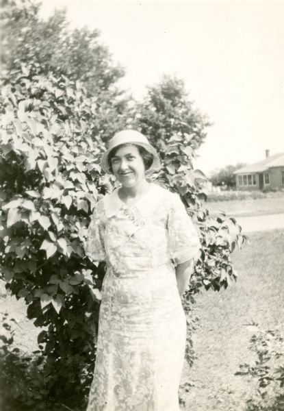 Mary Oungst, teacher at the Cloverland School, District No. 2, poses standing in front of a lilac bush. There is a house in the background.