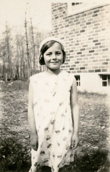 Phyllis Coggins, age 9, poses alongside the Cloverland School.  She is identified as the "Leader in English" among the school children.  She is wearing a summer dress and cap.