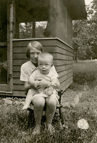 Helen Woodbury, age 12, a student at the Cloverland School, is pictured sitting in a small chair holding her baby brother, Jackie. They are posing just outside the door of a screened porch. She is described on the reverse of the photograph as the "Winner of several prizes in girls 4-H Club work. Good student and housekeeper."
