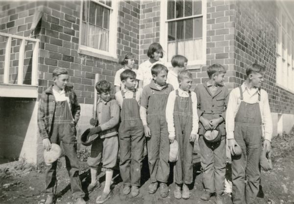 The kittenball team of Cloverland school, including three girls and seven boys, pose in front of the school. A boy in knickers holds a bat; another boy at far right holds a ball. The children have their heads turned to their left.  They were the town champions in 1932.
