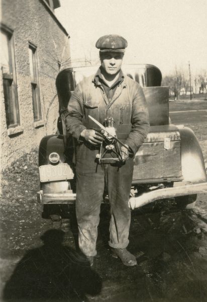 Harry Pederson poses while holding a model airplane motor which he built. He is standing outdoors in front of the rear of a car parked near a building.
