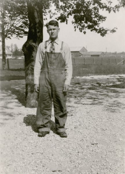 Alvin Sebald, 14, a student at Rhine Center School, poses in front of a tree. He is wearing bib overalls and a necktie. There is a barn and other farm buildings in the back ground. Alvin is described on the reverse of the photograph as "very strong; hard worker and player; boisterous; good student."