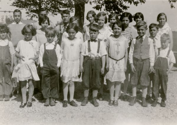 The students of the Rhine Center School, District No. 6, pose for their photograph in the schoolyard. The girls are wearing good dresses; the boys wear knickers or bib overalls. Some of the boys wear neckties.
