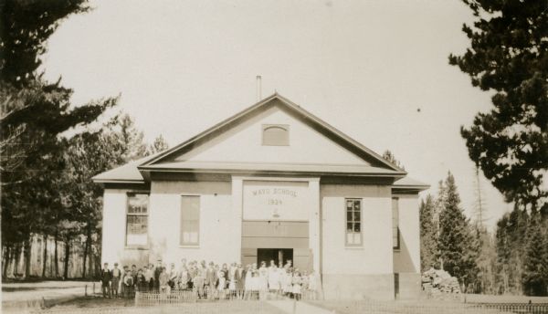 Students pose in front of the Mayo School, District No. 1. There are large pine trees around the one-story building and a low fence in front. A "Mayo School 1924" sign is above the door.