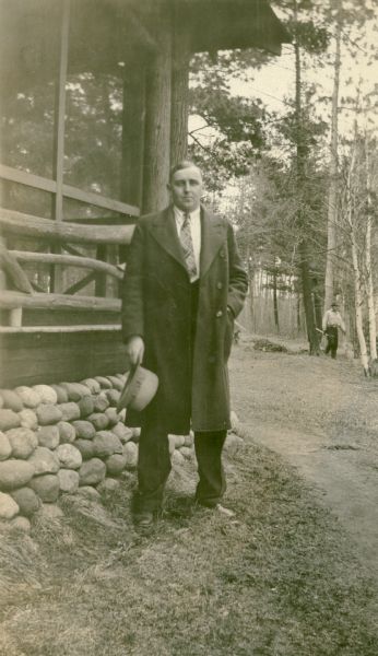 Francis Johnson, teacher at the Mayo School, District No. 1, standing beside a screened porch. The porch has a rustic log railing and stone foundation. There are pine and birch trees in the background. An unidentified man is walking in the background.