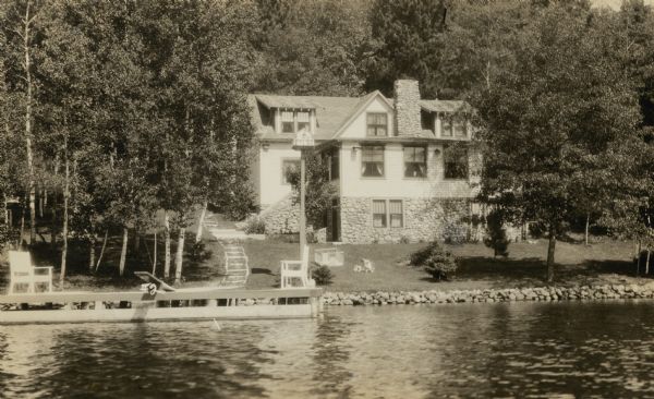 View from water of a child playing near a playpen on the lawn of a one and one-half story cottage on a slope along the shore of a lake. There is a stone seawall, chimney and foundation. A pier with benches and a diving board is in the foreground. A birdhouse for purple martins is mounted on a tall pole at the shoreline. On the reverse of the photograph is written "Private owned summer home four miles east of Eagle River on Cranberry Lake, Eagle Chain. Only native rock of this region was used in construction. Home owned by S. Ohnstein of Chicago."