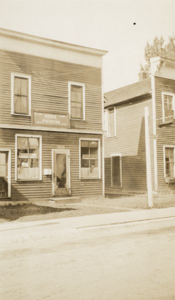 View from street of a two-story "boomtown" style building which was the residence of Francis Johnson, teacher at the Mayo School, District No. 1, Town of Washington, Vilas County. The first floor served as the business location of Rice Maid Photos. A sign above the door advertises Kodak Finishing, Printing, and 24 Hour Service. On the box to the left of the front door is written "Place Films Here." Johnson rented the building. A similar building stands to the right; decorative brackets adorn its cornice.