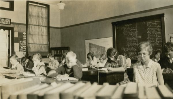 Students work at their desks in the Mayo School, District No. 1. A row of books forms the foreground; history lessons from the time of reconstruction after the Civil War are written on the blackboard.