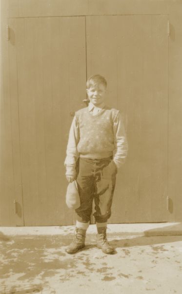 Gene Mayo, 14, poses in front of double doors holding his cap. He is a student at the Mayo School, District No. 1. On the reverse of the photograph is written, "Rated by Co. Nurse as the 'most physically fit' student in school. Exceedingly active — good student."