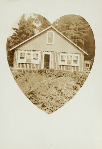 Pictured in a heart-shaped frame is a story and one-half house with vertical log siding on the lower floor with a stand of large pine trees in the background. It was the home of Charles Young, a student at the Mayo School, District No. 1. Charles was the "Leader in Mathematics" at the school. His father was Arthur Young.