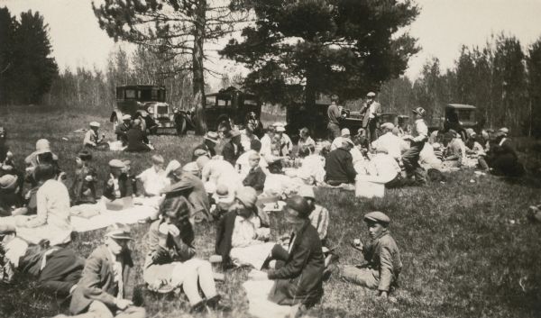 Students of the Mayo School, District No. 1, have gathered with their families for a picnic at a grove on the Deerskin River. Automobiles are parked under large pine trees in the background. On the reverse of the photograph is written: "4-H club meetings and other community gatherings held here when weather permits."