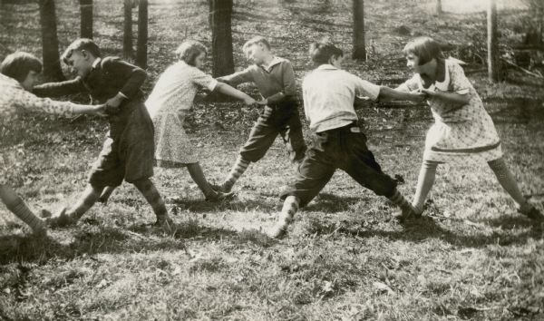 Three couples of school age children perform a folk dance outdoors in the grass. The boys wear knickers and patterned stockings; the girls are in dresses. They are students at the Fontana State Graded School.