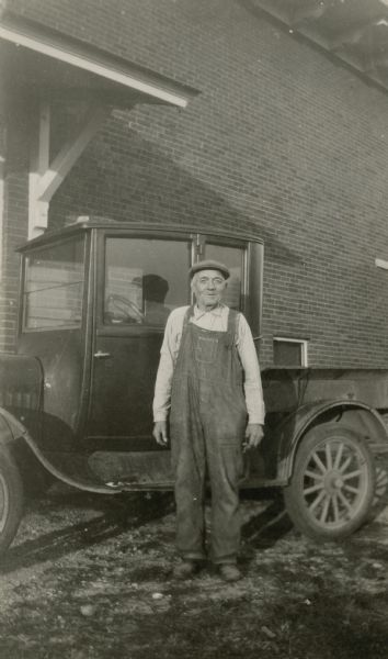 Emil Schwartzer, the janitor at Honey Creek School, District No. 1, poses in front of a pick up truck in front of the school building. He is wearing bib overalls and a cap.