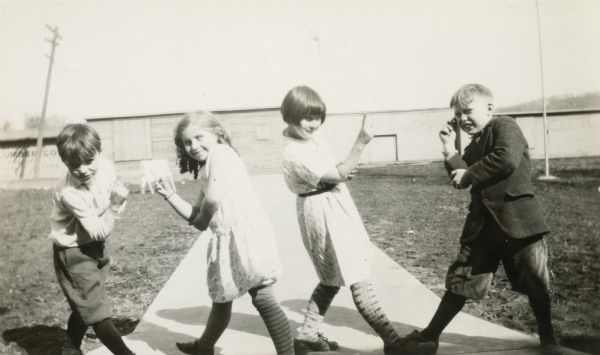 Four students of the Fontana State Graded School strike a pose while folk dancing outdoors. The boys are wearing knickers; the girls wear dresses and patterned stockings. There are several long, low wooden buildings in the background. A sign on one of the buildings identifies a lumber company.