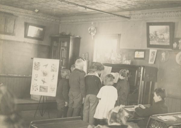 Several students have gathered around the piano for "Leisure Hour" at Star Center School, District No. 8, Bloomfield. Other students work at their desks. There is a poster of various birds on an easel at the front of the classroom. The room has a pressed tin ceiling and cornice.