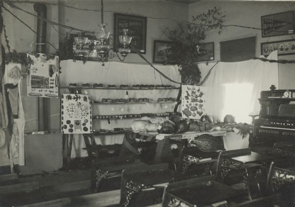 A display of autumn produce including corn, squash, cabbage and apples occupies a corner of the Centerville School. There are posters of animals, vegetables, and leaves. Framed prize banners from town spelling competitions hang on the walls. A three armed chandelier with oil lamps hangs from the ceiling and there is a parlor organ against the side wall. On the reverse of the photograph is written "School now closed, transporting to East Troy."