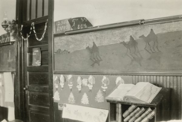 The Briggs School is decorated for Christmas with colored chalk drawings of the Nativity story on the blackboards. The section shown depicts the Three Magi. Cut-out depictions of Santa Claus and Christmas trees, as well as a drawing of Santa's sleigh and reindeer, hang beneath the blackboard. There is a paper chain over the door. A book stand holds a large dictionary with other books on the shelf below.