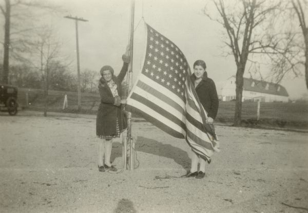 Two unidentified girls pose holding an American flag at base of a flagpole at Lauderdale Lake School. There is a large barn in the background and a car is parked on the left. On the reverse of the photograph is written "Typical of 'patriotic moment' in Morning Exercises in every school in Walworth County."