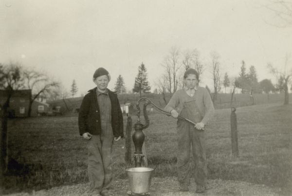 George Stallman and Walter Shinkus pose with a bucket at the water pump outside the Tibbets School. They are "getting water for the school."  There are houses, fields, and a hill in the background.