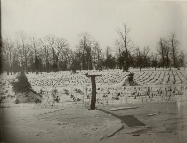 Identified as "A lunch counter for birds," a platform feeder has been mounted on a fence post behind Gardners Prairie School, District No. 7, Town of Spring Prairie. Beyond the fence, corn shocks and stubble protrude from drifted snow; there is wooded land in the background. A lone bird sits on the feeder which casts a long shadow on the snow.