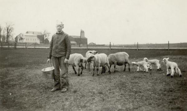 Warren Potter, holding a bucket, poses with his sheep and lambs.  There is a road in the background and a farmstead on the other side of the road.