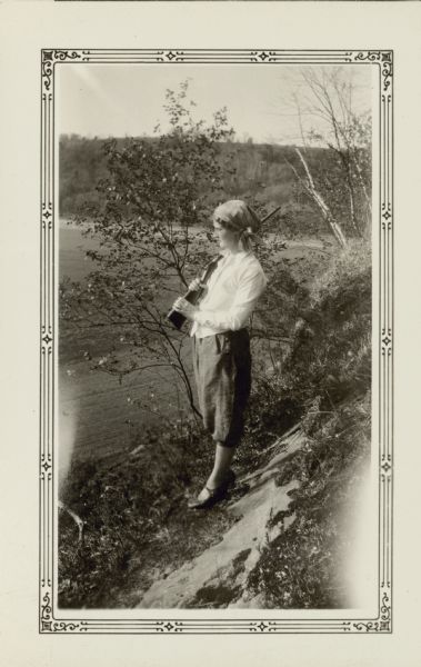 Selma Eidsmoe, wearing knickers and a head scarf, poses holding a gun over her shoulder. She is standing on a bluff above the Pecatonica River valley. She taught at the Farmers Grove School, District No. 3, Town of York and New Glarus.