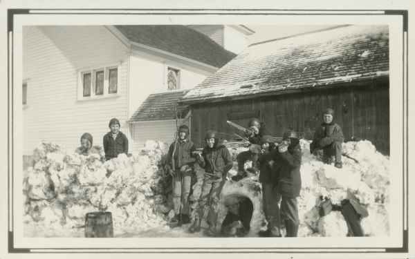 Winter scene with boys in winter coats and hats aiming toy stick guns while posing on an igloo and fort they have built from snow. A church building forms the backdrop. On the reverse of the photograph is written: "'Pupils Play' in their school yard, at home building 'Homes of Far Away Children' (Eskimo).  Gladys White - Teacher."