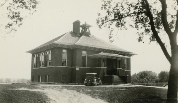 An automobile is parked near the entrance to Honey Creek School, District No. 1. The school is a sturdy brick structure with raised basement and hip roof. There is a large central chimney and a bell tower. Broad overhangs, horizontal banding in the brickwork and battered wall details on the porch suggest prairie style influence.