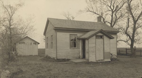 The rear wheel of a bicycle is visible beyond the small front porch of a small, deteriorating one room school.  The main portion of the school building is classical revival in design and proportion.   There is a second bicycle leaning against a tree on the left.  An outbuilding stands toward the rear of the school yard which is marked on the right by an old wooden fence.