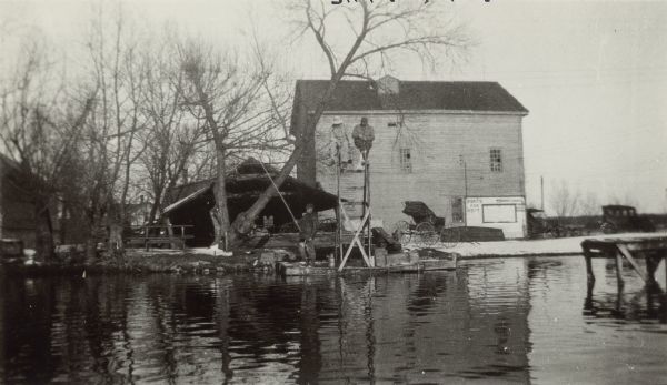 Two men fish from a diving platform while a third fishes from the shore at Jeffry's Mill. The tall, wood frame mill building is in the background, with an open carriage and two automobiles parked nearby. A sign on the mill advertises "Boats for Rent." There is a small amount of snow on the ground but no ice on the pond.