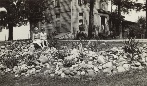 Two unidentified girls pose seated on a large stone in a rock garden on the lawn of the Charles E. Curver home. There are small clumps of flowers in bloom in the garden. The two-story wood frame house has a small recessed front porch with columns. There are window boxes at the two front first-story windows. Curver's daughter Harriet was the teacher at Hillcrest School, District No. 6, Towns of Waukesha, Pewaukee, Brookfield and New Berlin. The house was built by Isaac Pigeon Walker, United States Senator from Wisconsin from 1848 until 1855.