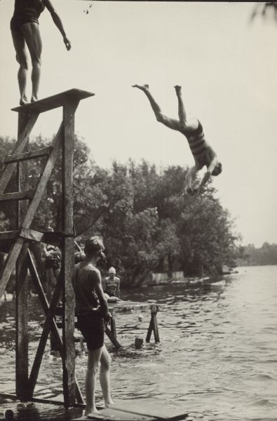 A diver is caught mid-air as another prepares to dive from a diving platform on the shore of the mill pond at Jeffry's Mill at Eagleville. A boy watches from the base of the platform; others sit or stand on a pier in the water.