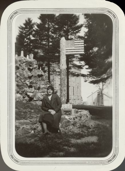 Miss Barbara Kiechler, identified as a 4-H Club worker in Coon Valley, posing on a low wall surrounding the Grotto of the Holy Family on the grounds of Villa St. Joseph, a convent. A mosaic stone flagpole and flag stand behind her. In the background is the stone convent building, and beyond that, a barn and silo. The convent was established by the Franciscan Sisters of Perpetual Adoration in 1898. The grotto was built 1925-1930 by Father Paul Dobberstein.