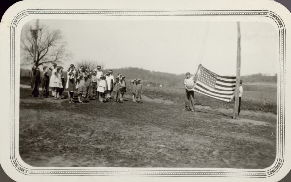 Two boys display the American flag before raising it on a rustic flag pole in the school yard of the Consolidated School, District No. 4. The other students stand and salute. There is a farmstead in the background.