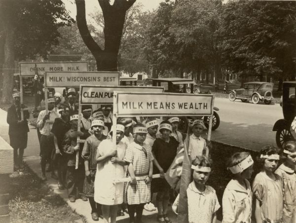 School children are lined up four abreast on the sidewalk; alternate rows carry signs or flags promoting milk. Each child also wears a head band with "HEALTH" printed on it. Cars are parked along both sides of the shaded street.