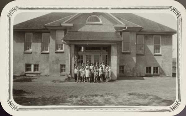 Children stand in a front of the Consolidated School, District No. 4. The stucco building has an elevated basement and large front porch.
