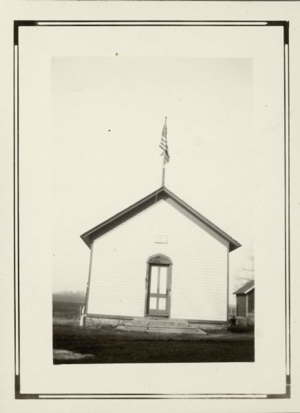 A simple sign over the front door identifies the one-room Fairbanks School. A flag hangs from a pole mounted on the roof. The top of the door is ornamented with a pointed arch. There is an outhouse on the right.