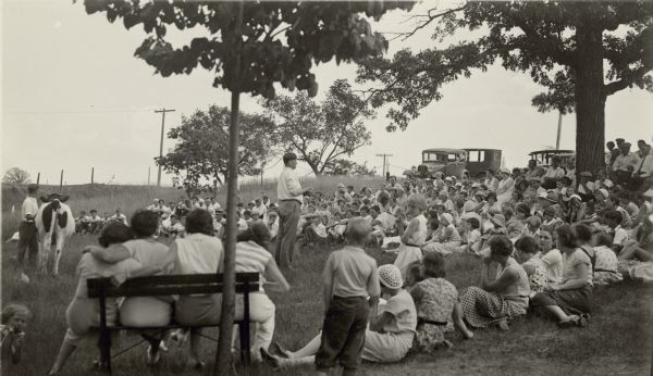 J.C. Nisbet (center), an editor of <i>Hoard's Dairyman</i> magazine, addresses a crowd of men, women and children at the Walworth County 4-H Club Picnic at Booth Lake Park. A man holds a Holstein calf at left. Automobiles are parked behind the crowd.