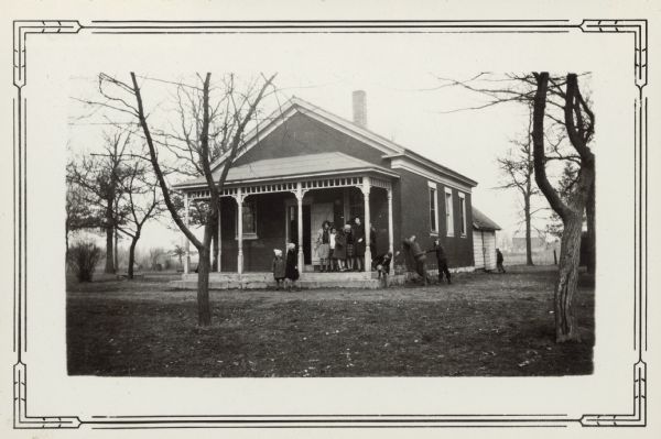 Students of the Battle Creek School, Joint District No. 4, have gathered outside the school building for their picture. The girls are on or near the front porch; two boys are at the side of the school while another boy runs toward the rear. The school is a small brick structure with a front porch and wooden rear addition. There is a barn in the background.