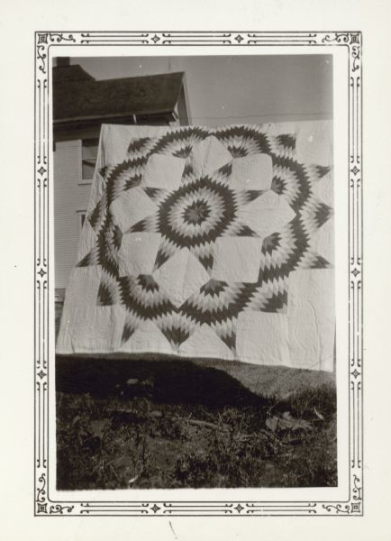 A quilt made by Elda Strahm, a student at Farmer's Grove School, town of York and New Glarus, is displayed hanging outdoors on a clothes line.  There is a large house in the background.