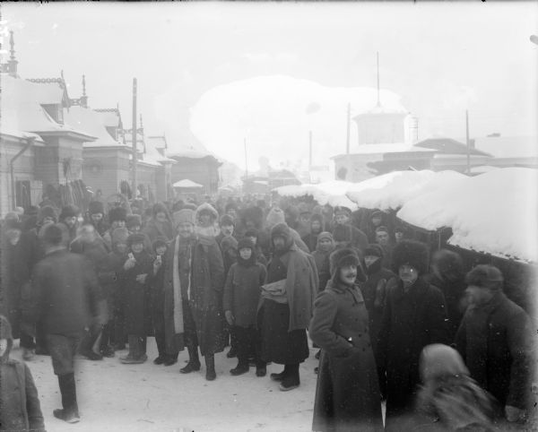 Slightly elevated view of a group of men, women, and children standing outdoors and looking towards the camera. They are probably attending the market in Arkhangelsk, Russia. Amongst the group of people there appear to be a few soldiers, one of which is wearing a cap with the emblem for the British grenadiers.