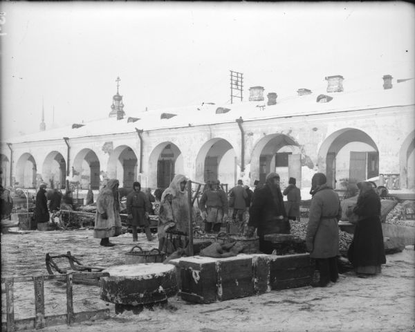 People wearing thick coats and hats, some made of fur, are standing at an outside market, possibly in Arkhangelsk, Russia. In the background is a long building with a row of archways.