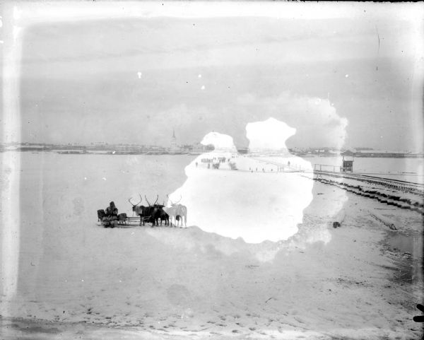 View across snow of a man driving a sled with a team of reindeer. There are railroad tracks on the right that run towards a town in the distance. A number of people are traveling on the ice near the railroad tracks near the far shoreline.