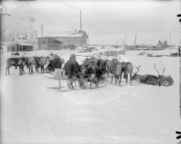 Two men and a young boy pose on sleds near their teams of reindeer in front of a house and its outbuildings. In the background is a large building with onion domes. In the background on the right men work near stacks of logs.