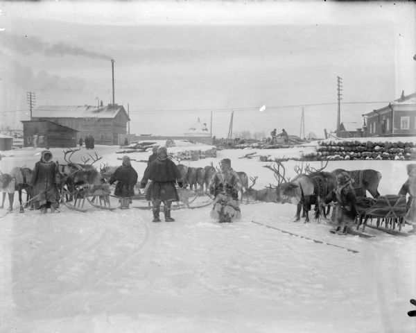 A family of Laplanders with their reindeer sled teams. In the background are a house with its outhouses, and a group of people to the left of center, as well as two men sawing a piece of timber.