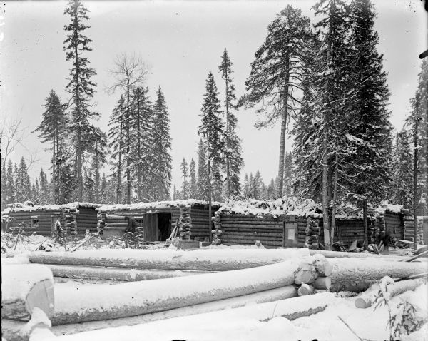 View across snow-covered logs of billets of the 310th U.S. Army Corps of Engineers near Arkhangelsk, Russia surrounded by pine trees. There are pine boughs on the flat roofs of the billets. Near the middle barrack is a man working outdoors, and another man is standing in an open doorway looking toward the camera.